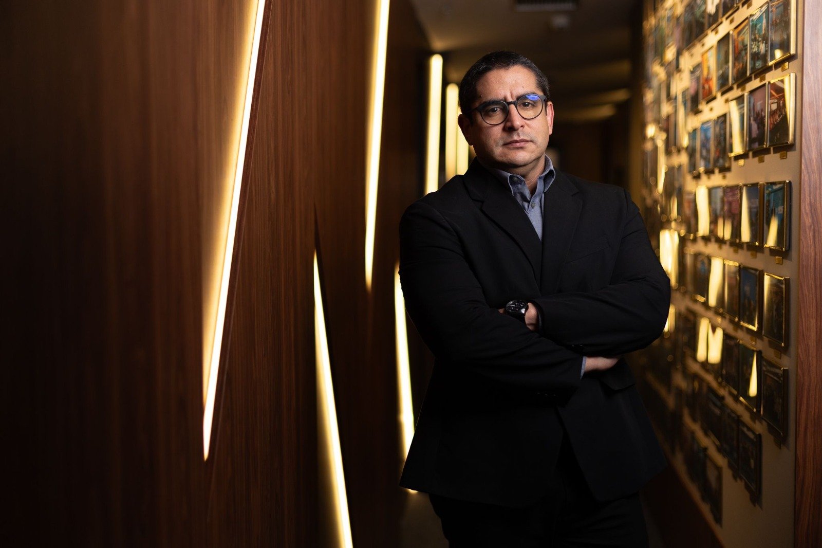 Dr. Romulo Victor, de terno preto e braços cruzados, em um corredor de madeira com luzes